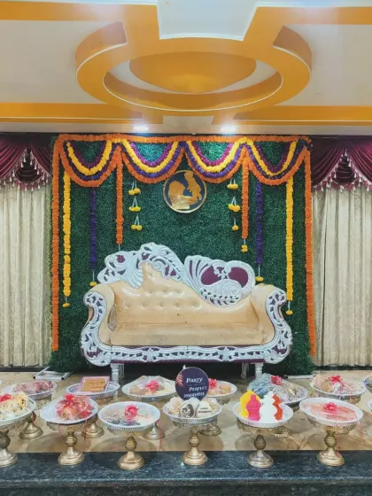 A wide view of a traditional baby shower (Seemantham) setup, showing the ornate seating against a green grass and floral backdrop. The stage is filled with numerous offering trays, showcasing the scale and completeness of the ceremony decor I provide.