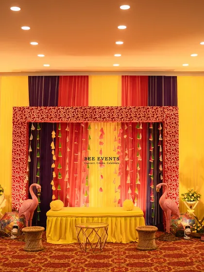 A colorful and quirky Mehendi stage. The backdrop features vibrant drapes in yellow, pink, and purple, with fun elements like flamingo props and tassel hangings, creating a lively and celebratory mood.