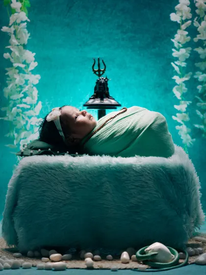 A divine portrait celebrating new life, this Maha Shivratri theme features a peacefully sleeping newborn swaddled in soft green fabric. The setup includes a miniature Trishul and Shivling against a serene turquoise backdrop, creating a truly unique and spiritual memory.