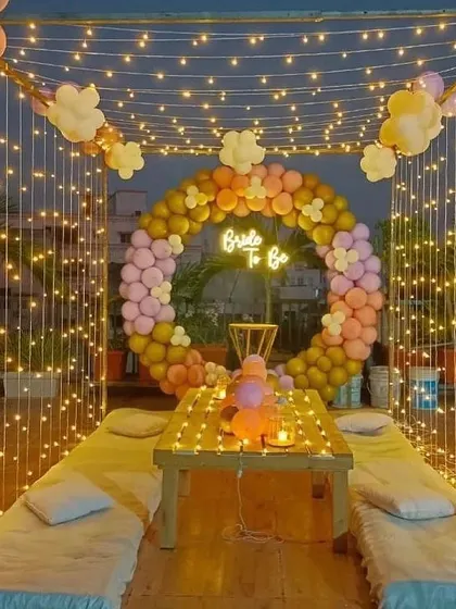 I created this magical bride-to-be party on a terrace, perfect for an evening celebration. The setup includes a canopy of fairy lights, low seating with cushions, and a circular balloon arch in pastel shades. A 'Bride to Be' neon sign serves as the centerpiece for photos.