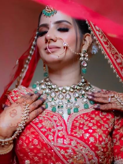 A close-up of the bride adjusting her stunning emerald and polki necklace, with the veil softly framing her face. This shot focuses on the luxurious bridal jewelry.