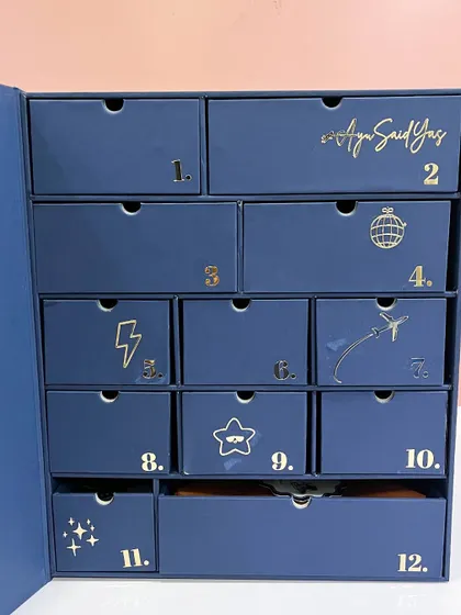 The front of the advent calendar, showing the 12 numbered drawers, each decorated with a unique gold foil icon representing the gift inside.