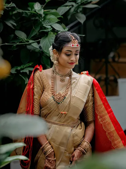A quiet moment captured in a garden. The bride, in her beautiful gold and red saree, looks down with a gentle smile, a picture of grace and tranquility.