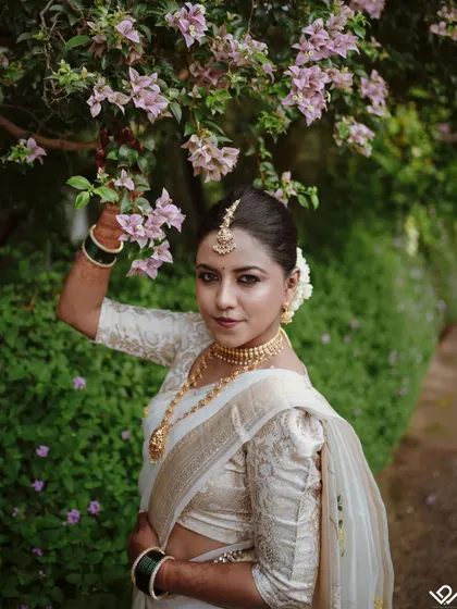 A beautiful outdoor bridal portrait, with the bride posing under a bougainvillea tree, perfectly complementing her white and gold saree.