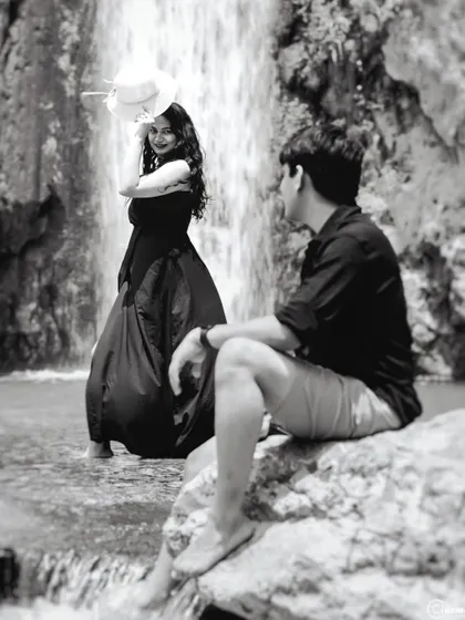 A black and white photo capturing a candid interaction by a waterfall. The composition creates a story, with the man looking on as the woman stands poised and elegant against the flowing water.