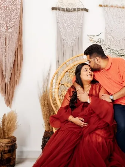 A loving embrace in the studio. The partner kisses the mother-to-be's forehead as she sits in a wicker chair, surrounded by a cozy, rustic setup.