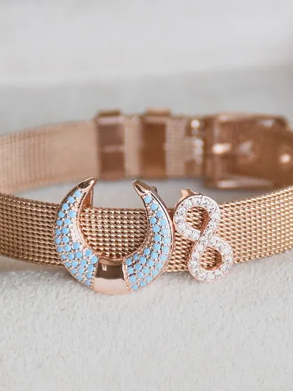 A close-up of this stunning rose gold watch belt. It features a turquoise-studded crescent moon charm paired with a sparkling infinity symbol.
