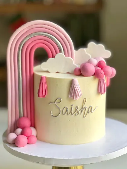 A boho-chic rainbow cake in shades of pink and silver. The fondant rainbow is accented with tassels and fluffy clouds, creating a trendy and stylish look for a birthday celebration.