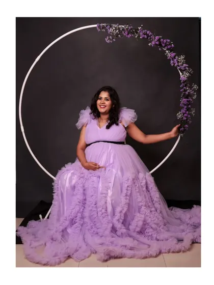 The expecting mother seated with the floral ring prop, showcasing the full, beautiful spread of the lavender ruffle gown.