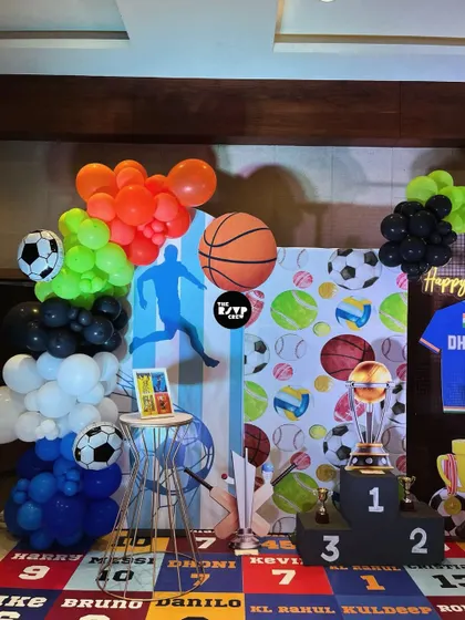 This sports party setup includes a winners' podium, a basketball-themed balloon arrangement, and a backdrop filled with different sports balls.