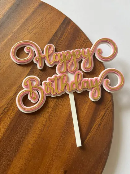 A whimsical layered paper "Happy Birthday" topper in shades of pink and gold. The swirly font adds a touch of fun and elegance.
