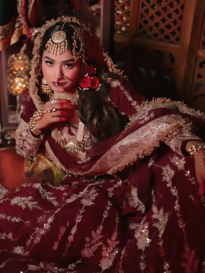 A full-body shot of the bride seated gracefully. The rich maroon lehenga spreads around her, showcasing its heavy embroidery and creating a truly royal portrait.