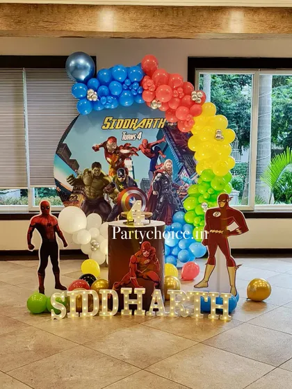 A powerful superhero-themed birthday party featuring the Avengers. The decor includes a dynamic backdrop, character cutouts of Spider-Man and The Flash, and a colorful balloon arch.