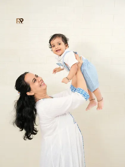 A heartwarming portrait of a mother lifting her smiling baby. The simple white brick background keeps the focus on their beautiful interaction and loving bond.