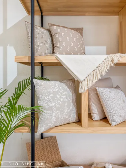 A close-up of the custom shelving, showing the beautiful textures and patterns of FA Home's cushions. The combination of light wood and black metal supports provides a modern yet warm framework for the products.