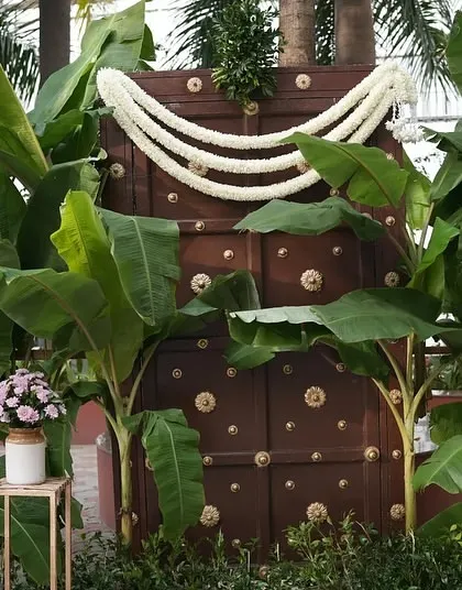 A traditional South Indian photo backdrop featuring an antique wooden door, banana plants, and jasmine garlands.