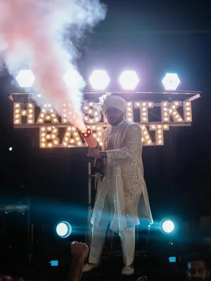 The groom makes a grand entrance for his baraat, using a smoke gun to amp up the energy. The custom 'Harshit ki Baraat' sign adds a personal and fun touch.