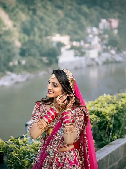 The bride adjusts her earring, with the beautiful Ganga river and Rishikesh landscape creating a breathtaking backdrop.