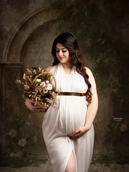 A gentle and beautiful portrait of a mom-to-be in a white gown, holding a basket of flowers. The dark, painterly background makes her stand out, creating a classic, artistic feel.