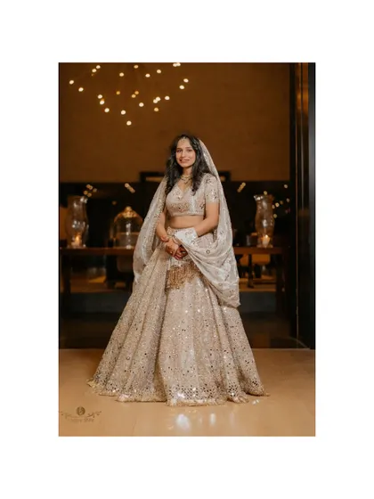 An elegant full-length portrait of the bride in her ivory lehenga, standing in a grand, warmly lit hall.
