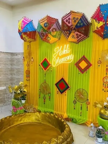 An indoor Haldi ceremony corner with a large golden 'urli' and a backdrop of green and yellow drapes, decorated with umbrellas and a neon "Haldi Ceremony" sign.