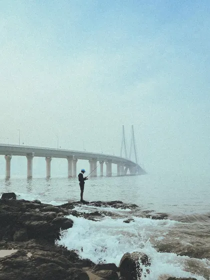 A lone fisherman stands on the rocks against the foggy backdrop of the Bandra-Worli Sea Link.