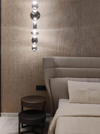 A detail of a bedroom corner, focusing on the interplay of form, texture, and light. A pendant light with stacked glass elements hangs beside a high-back upholstered bed, against a backdrop of vertically textured wallpaper.