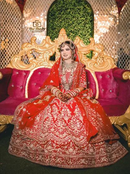 A royal bridal portrait. The bride is seated on a grand, ornate sofa, showcasing her magnificent red lehenga in all its glory.