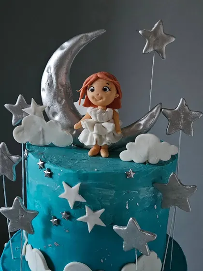A close-up of the top tier of the moon-themed cake. This view highlights the handcrafted girl figurine sitting on the shimmering silver moon, surrounded by edible clouds and stars.