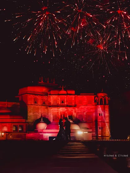 A romantic silhouette of the couple against the red-lit Fairmont Jaipur palace, with fireworks overhead. A truly magical and iconic shot.