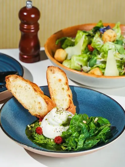A fresh burrata salad with pesto, greens, and toasted bread, served in a beautiful blue ceramic bowl. The bright, hard light creates a clean and contemporary feel, perfect for a modern cafe menu.