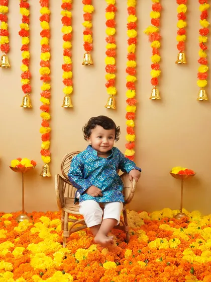 A little boy dressed in a traditional blue kurta for a Ganesh Chaturthi themed shoot. The backdrop of marigold flowers and bells creates a vibrant and festive atmosphere.