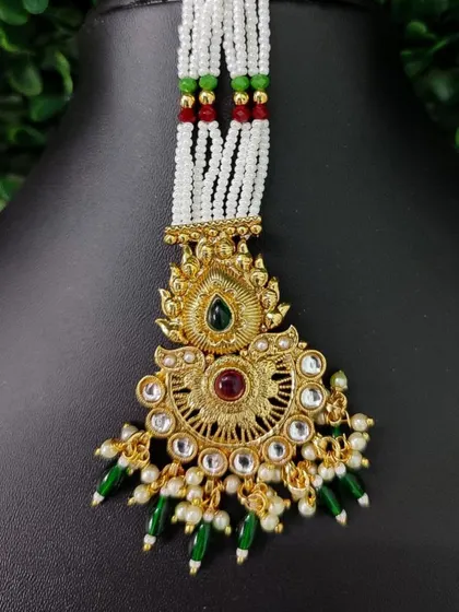 A close-up of a maang tikka with a traditional design, featuring red and green stones and green bead drops, attached to white pearl strands.