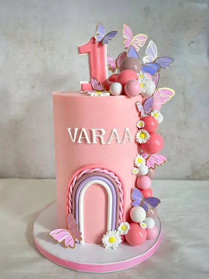A pretty first birthday cake for Varaa, featuring a tall pink design with a cascade of butterflies, bubbles, and a delicate rainbow.