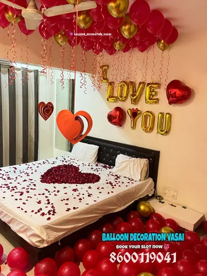 A full view of a bedroom filled with red and gold balloons on the floor and ceiling. The bed is the centerpiece, with a large rose petal heart and "I LOVE YOU" message.