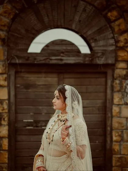 A classic side-profile portrait of the bride in her white lehenga, set against a rustic stone archway.