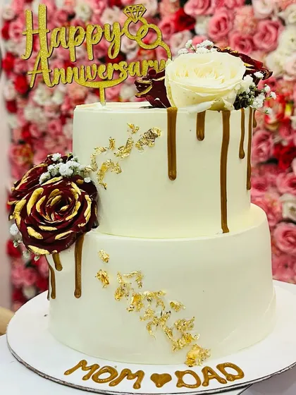 A two-tiered anniversary cake for 'Mom & Dad'. The design features a caramel drip, gold leaf accents, and beautiful red and white roses.