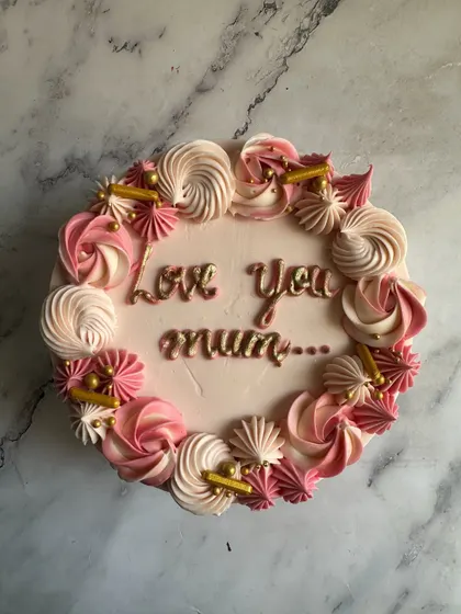 A sweet and simple cake for Mom, with "Love You Mum" written in sparkling gold. The border is decorated with a wreath of pink and white piped meringues and sprinkles.