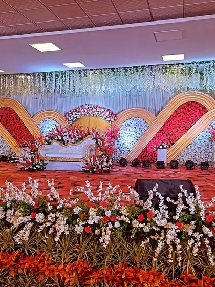 A grand indoor reception stage with a multi-layered backdrop of red and white floral panels, creating a bold, artistic statement.