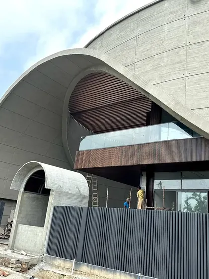 Another view of the family home in Haryana, showing the interplay between the sweeping concrete arch, the warm wood cladding, and the dark vertical fins of the fence. The design creates a dramatic entrance while maintaining a sense of privacy.
