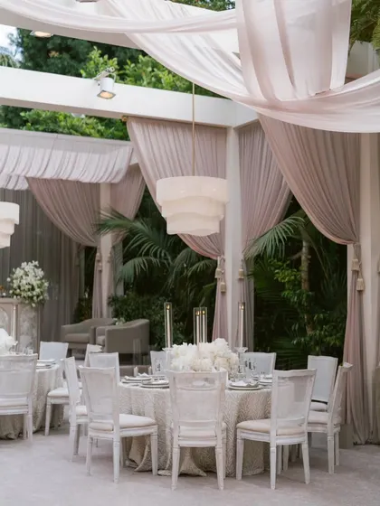 An elegant dining area draped in soft, neutral fabrics. The modern tiered chandeliers and all-white table settings create a serene, sophisticated, and cohesive design.