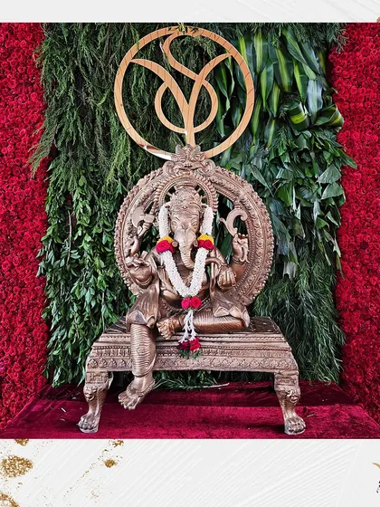 A modern and elegant Ganesha setup. The brass idol is placed against a backdrop of vibrant red flowers and green foliage, with a custom monogram above, blending tradition with contemporary style.