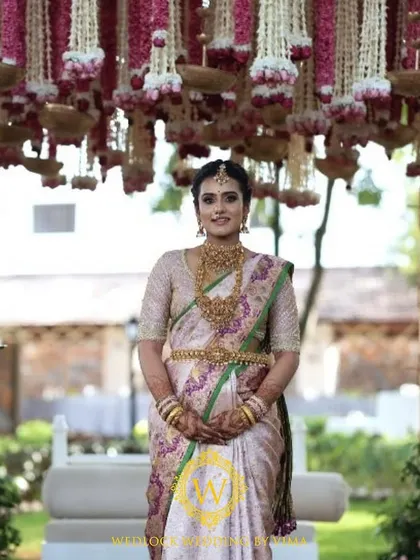 A beautiful bride standing before a traditional mandap decorated with pink and white floral tassels. The hanging brass urlis and rose drops add a touch of classic elegance to the South Indian ceremony.