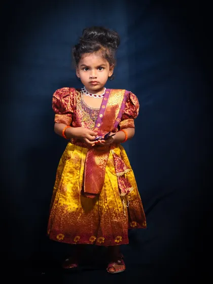 A full-length studio portrait of a young girl in a beautiful traditional dress. The dark, classic backdrop makes her colorful outfit pop, creating a formal and elegant look.