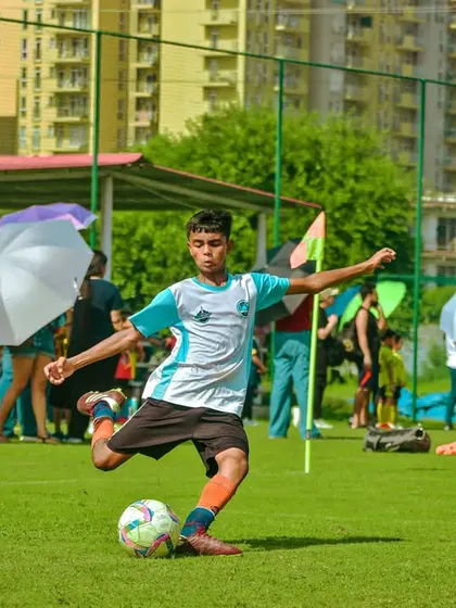 A player strikes the ball under the watchful eye of parents holding umbrellas on a sunny day.
