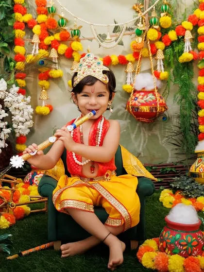 A little Krishna plays the flute in a lush, green setup decorated with marigolds and peacocks.