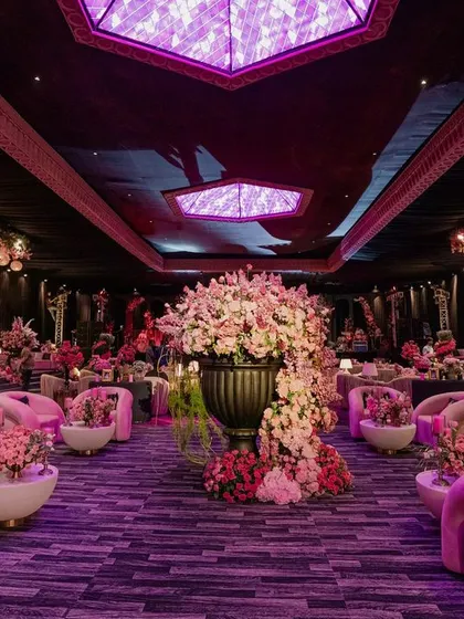 A wide view of the event space, showing the tiered lounge areas with their plush pink seating. A massive floral urn stands in the center, and the ceiling features dramatic purple lighting.