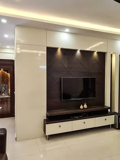 A contemporary living room featuring a sleek TV unit with a dark wood geometric panel and high-gloss white drawers. The layered false ceiling with cove lighting provides soft, ambient illumination, while a traditional pooja unit is visible in the background.