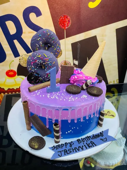 A 2.5kg drip cake in purple and blue, loaded with donuts, an ice cream cone, and chocolate bars for a first birthday.