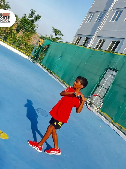 A young athlete in red practices his forehand on the blue hard court during a sunny day at camp.